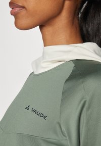 Indumento in tessuto leggero verde con collarino bianco, caratterizzato da un logo nero visibile, una texture liscia e un design semplice e funzionale.