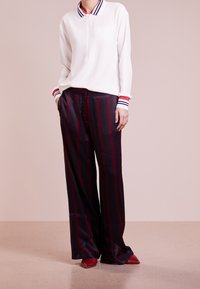 Polo bianco a maniche lunghe con polsini a righe, abbinato a pantaloni a palazzo con strisce verticali bordeaux e blu navy. Scarpe rosse a punta.