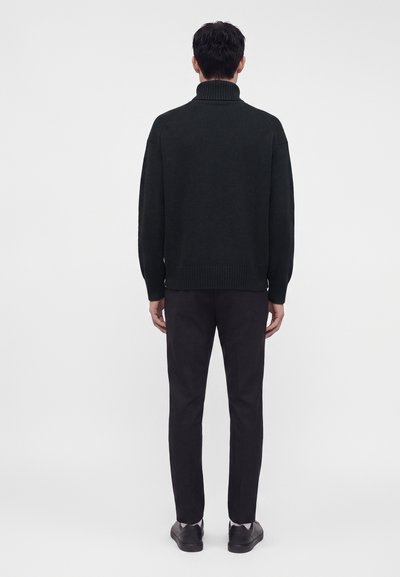 Homme portant un pull à col roulé noir, un pantalon noir et des chaussures noires, debout, tourné dos à un fond clair uni.