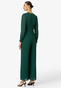 Groene jumpsuit met lange doorzichtige mouwen, brede benen en een ronde halslijn. Achterkant met ritsluiting en zichtbare knoopaccenten op de manchet.