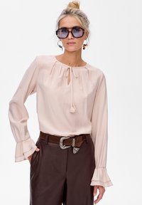 Jasnoróżowa bluzka z długimi rękawami, dekoltem w kształcie łezki i falbaniastymi mankietami, zestawiona z brązowymi skórzanymi spodniami i ozdobnym paskiem.