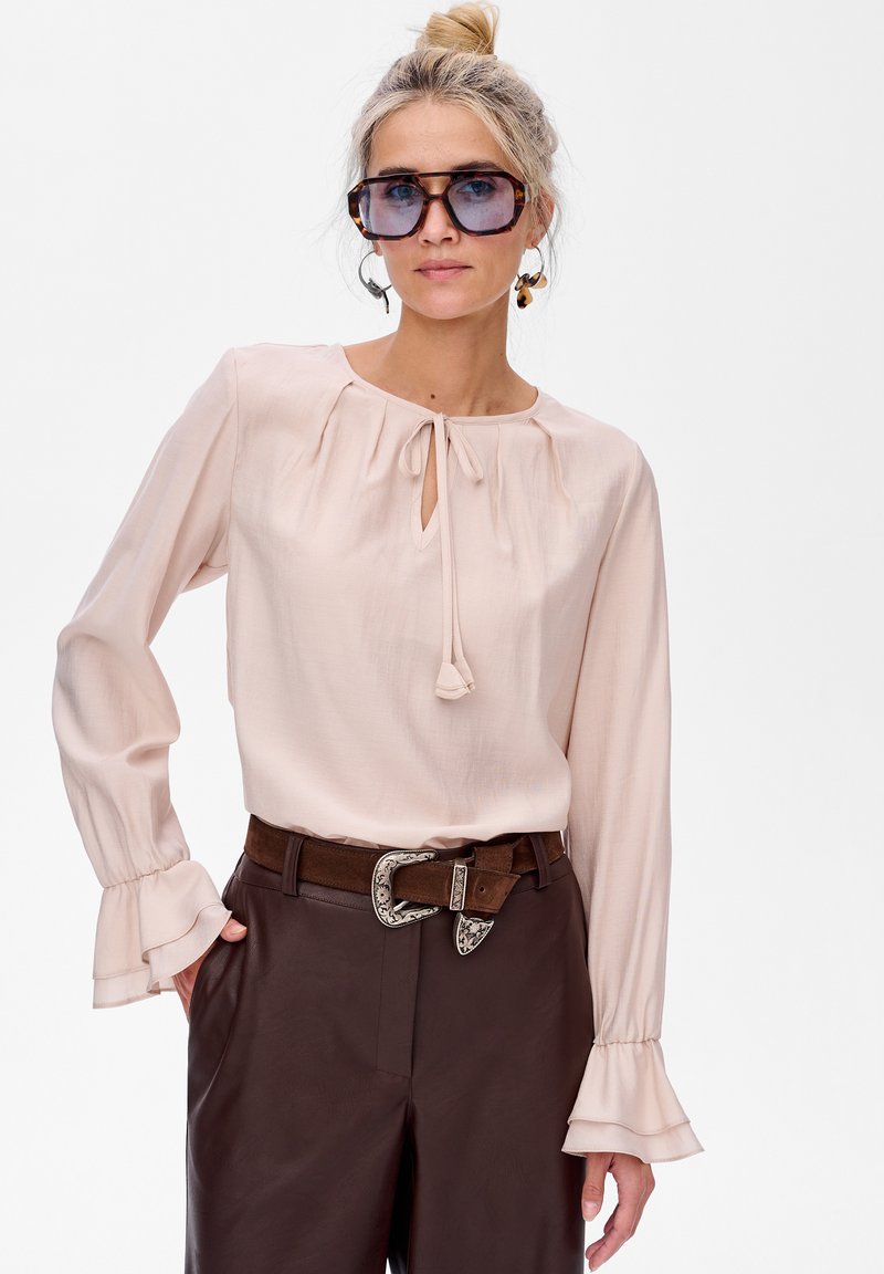 Jasnoróżowa bluzka z długimi rękawami, dekoltem w kształcie łezki i falbaniastymi mankietami, zestawiona z brązowymi skórzanymi spodniami i ozdobnym paskiem.