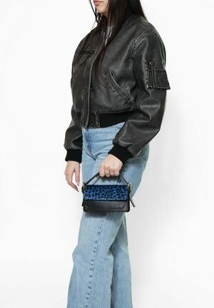 Personne portant une veste aviateur en cuir noir et un jean bleu clair, tenant un petit sac à main noir avec un rabat imprimé léopard bleu.