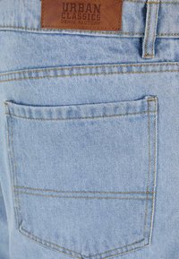 Tejido de mezclilla azul claro con un acabado texturizado, que presenta un bolsillo trasero y una etiqueta de cuero marrón con "Urban Classics Denim Factory."