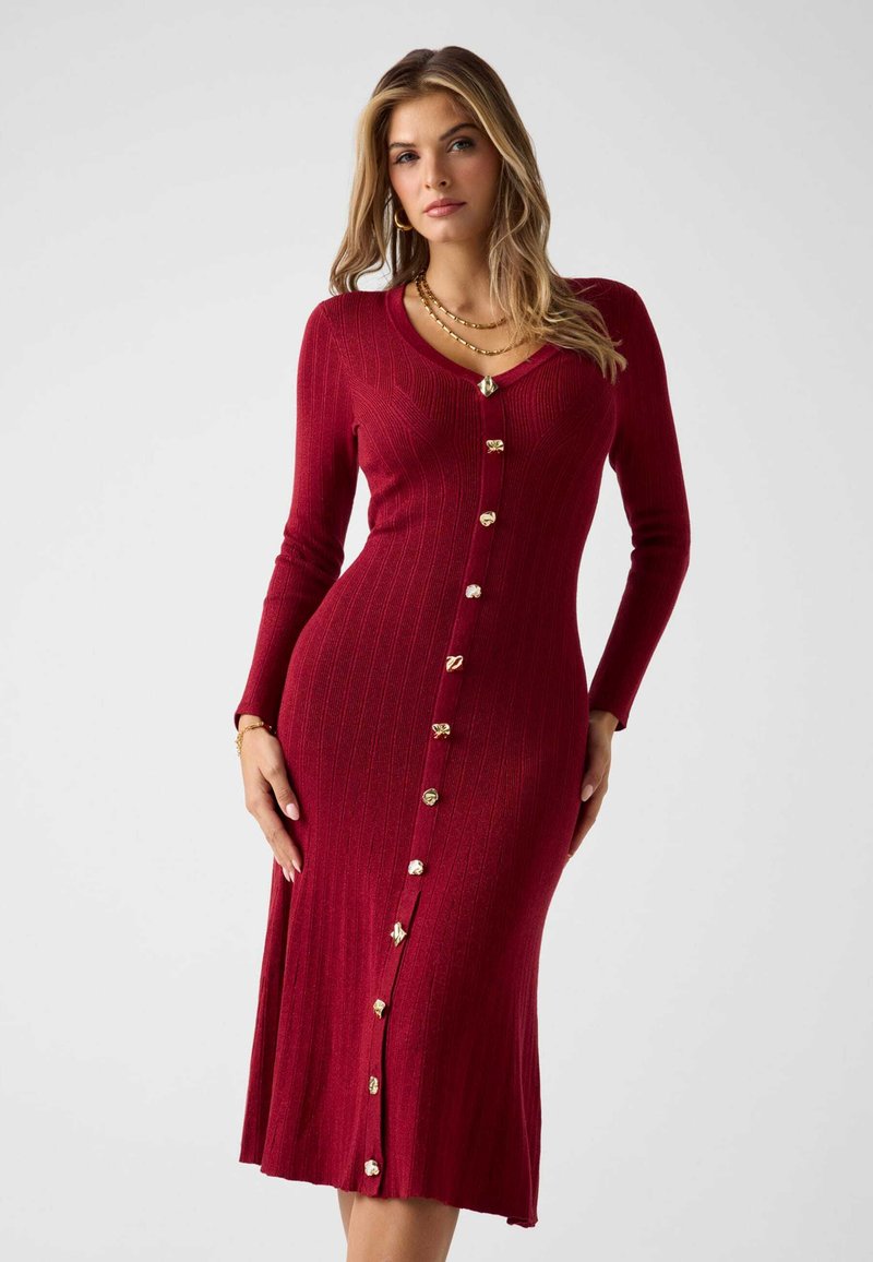 Geripptes rotes Kleid mit langen Ärmeln, V-Ausschnitt und goldfarbenen Knöpfen entlang der Vorderseite. Knielang mit leicht ausgestelltem Saum.