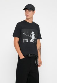 T-shirt de algodão preto com uma estampa gráfica de um baterista vestido com um fato de stormtrooper. Combinado com calças largas pretas e um boné preto.