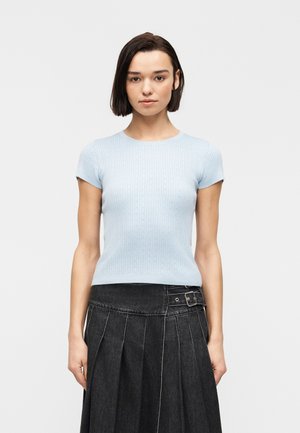 Jeune femme aux cheveux lisses et foncés portant un haut côtelé bleu clair à manches courtes et une jupe plissée noire taille haute avec ceinture.