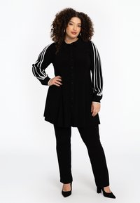 Tunique noire avec fermeture à boutons, dotée de manches longues avec des rayures blanches. Associée à un pantalon noir et des chaussures à talons.