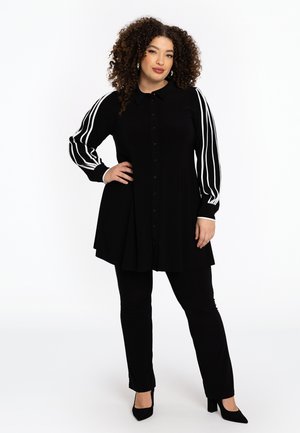 Tunique noire avec fermeture à boutons, dotée de manches longues avec des rayures blanches. Associée à un pantalon noir et des chaussures à talons.