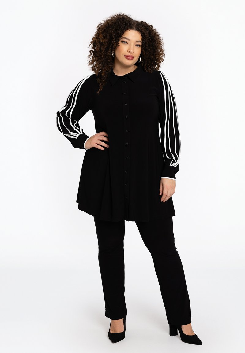 Tunique noire avec fermeture à boutons, dotée de manches longues avec des rayures blanches. Associée à un pantalon noir et des chaussures à talons.