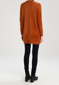 Lång orange fluffig cardigan med avslappnad passform, rund halsringning och en något oversized look, parat med mörka skinny jeans och ankelstövlar.