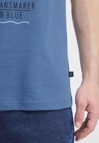 Tricou din bumbac albastru cu textul "TRANSMAKER IN BLUE", mâneci scurte și etichetă pe lateral. Purtați cu blugi din denim albastru. Textură netedă.