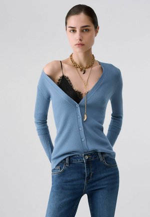 Jeune personne portant un cardigan bleu boutonné tombant des épaules sur un top en dentelle noire, un jean bleu et des colliers dorés superposés, posant devant un fond uni.