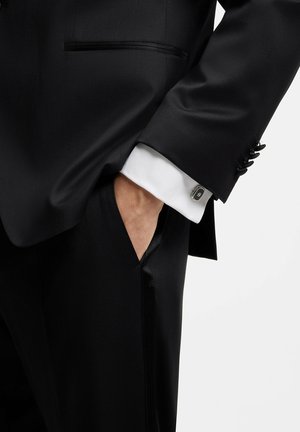 Mand iført sort smoking med hvide manchetter og sølv manchetknapper, hånd delvist i lommen mod ensfarvet baggrund.