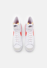 Bílé kožené tenisky Nike ve vysokém provedení s oranžovými akcenty, gumovými podrážkami a texturovanými šedými semišovými špičkami. Šněrovací design s logem.