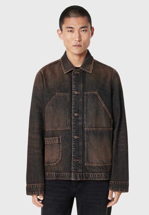 Ung mand iført en falmet sort denimjakke med store forlommer og knaplukning, stående mod en ensfarvet lys baggrund.