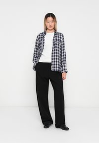 Jonge vrouw staat tegen een effen witte achtergrond, gekleed in zwarte broek, wit shirt en een ongeknopte blauw-wit geruite blouse, met handen in de zakken.