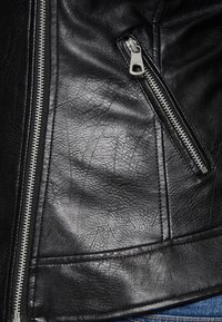 Svart fusk läderjacka med en texturerad yta, silverdragkedjedetalj på sidan och synliga stygnaccenter längs sömmarna.