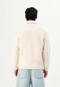 Man with dark hair stands facing away wearing a cream knit sweater and light blue denim jeans against a white background.