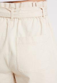 Pantalons beiges à taille élastique avec des détails froncés, comportant une poche arrière, fabriqués dans un tissu en coton texturé.