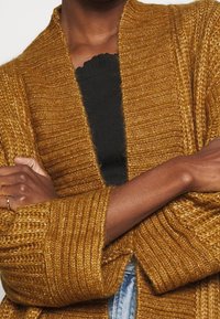 Senapsgul stickad cardigan med ribbad textur, öppen fram och vida ärmar. Bärs över en svart topp som har framträdande sömdetaljer.