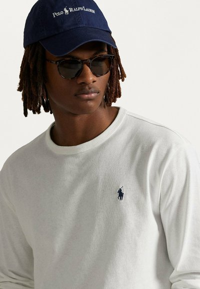 T-shirt bianco a maniche lunghe con logo blu navy sul lato sinistro del petto, abbinato a occhiali da sole scuri e un cappellino blu navy con branding bianco.