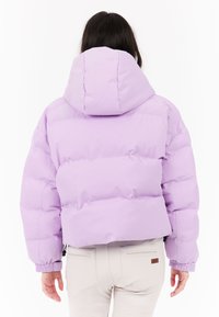 Žena s dlhými tmavými vlasmi, oblečená v svetlo fialovej prešívanej bunde s kapucňou a krémových nohaviciach, zobrazená zozadu na bielom pozadí.