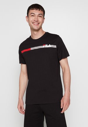 Homme souriant portant un t-shirt noir FILA avec des rayures horizontales rouges et grises, debout devant un fond gris clair uni.