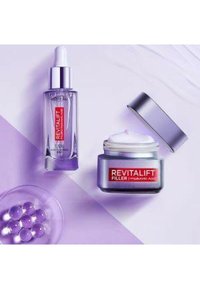 Sérum Revitalift dans un flacon en verre transparent avec pipette, et remplissage Revitalift dans un pot argenté et violet avec une crème blanche. Fond violet, accents brillants.