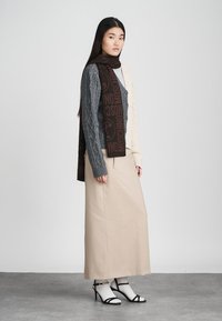 Gehaakte cardigan in grijs en crème, lange beige rok en zwarte sandalen met hak en bandjes, met kabelpatronen en logo-detail op de sjaal.