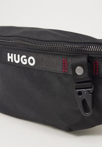 Juodas audinio juosmens krepšys su išraiškingu baltu "HUGO" logotipu, užtrauktuku ir juodu apdaila. Juostos akcentams naudojami raudoni siuvinėjimai.