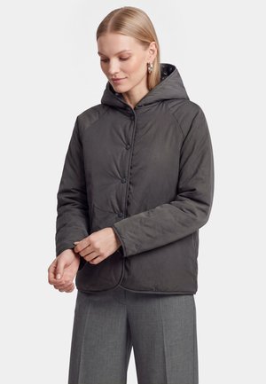 Femme portant une veste à capuche gris foncé avec fermeture par boutons et pantalon gris, ajustant sa manche, sur un fond uni.