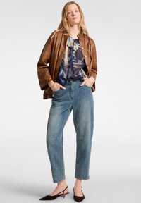 Chaqueta de cuero marrón sobre una blusa azul con estampado y volantes, combinada con unos jeans de mezclilla azul claro y zapatos de tacón marrón oscuro.