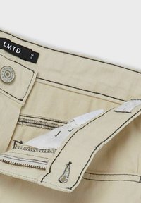 Béžové kalhoty s černým prošíváním, vybavené zipovým zapínáním, knoflíkovou légou a štítkem s nápisem "LMTD." Otevřené pro zobrazení detailu vnitřní kapsy.
