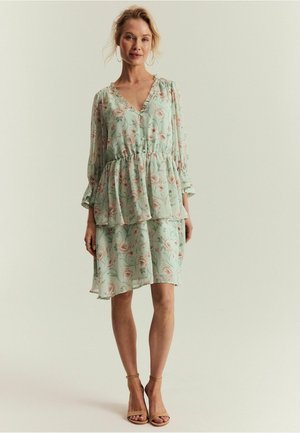 Kvinde stående iført en lysegrøn floral lagdelt kjole med lange ærmer og nøgenfarvede høje hæle mod en ensfarvet baggrund.