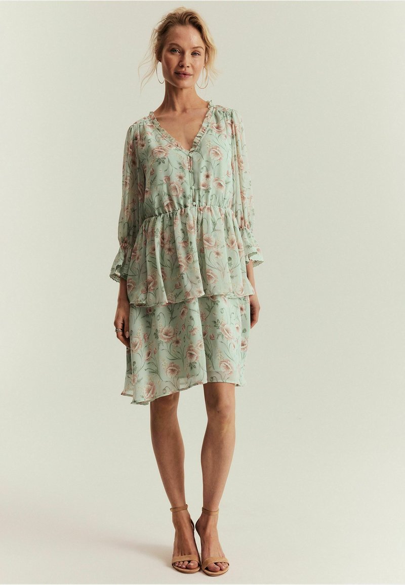 Frau steht und trägt ein hellgrünes, florales, geschichtetes Kleid mit langen Ärmeln und nude-farbene Highheel-Sandalen vor einem einfarbigen Hintergrund.