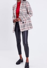 Blazer en tweed avec un motif à carreaux rouge, noir et blanc, fermeture à un bouton, associé à un pantalon en simili cuir noir et des mocassins noirs.