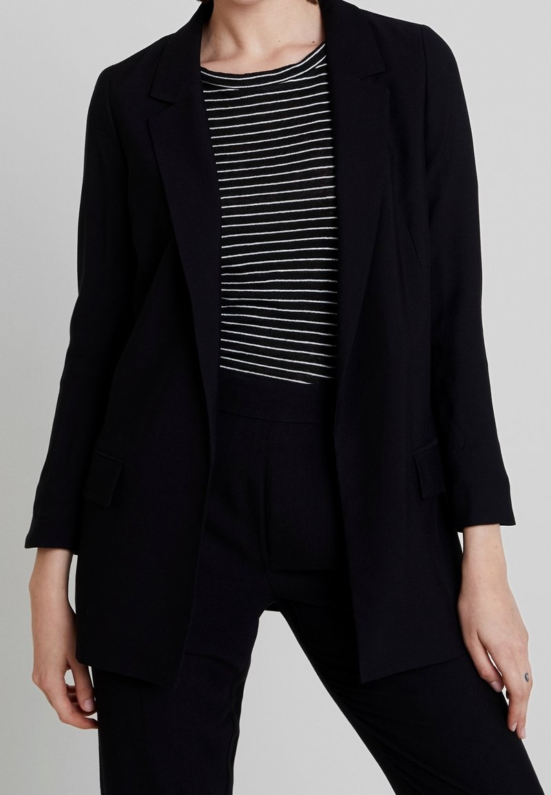 Personne portant un blazer noir sur une chemise rayée noir et blanc avec un pantalon noir, debout devant un fond gris uni.