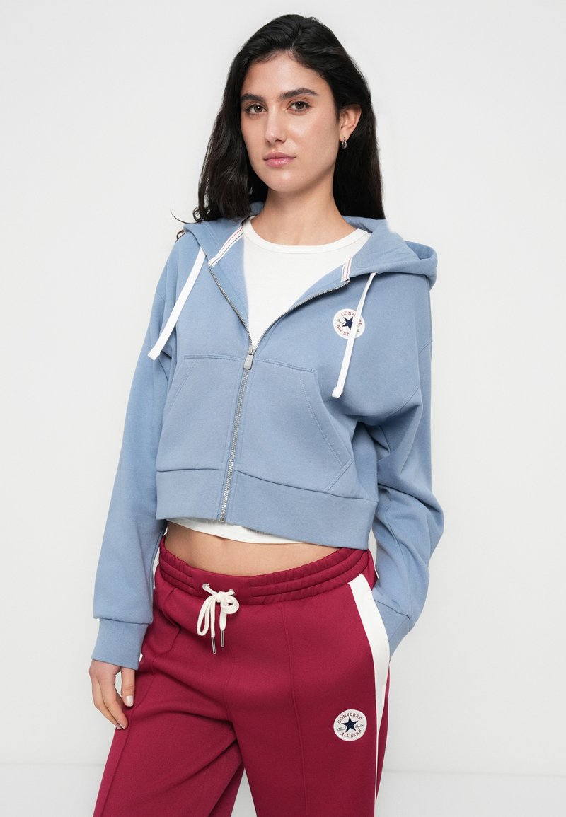 Sweat à capuche bleu recadré avec une fermeture éclair à l'avant, des cordons blancs et un patch logo. Associé à un pantalon de survêtement bordeaux avec des bandes blanches sur les côtés.