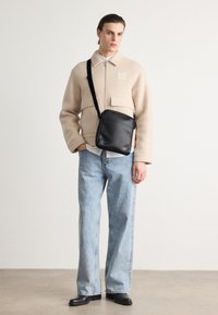 Bolso cruzado de cuero negro con una textura suave, bolsillo frontal con cremallera y correa ajustable, llevado sobre una chaqueta de forro polar beige y jeans azules.