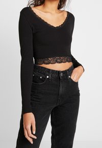 Frau in einem schwarzen langärmeligen Crop-Top mit Spitze und hochgeschnittenen schwarzen Jeans, Hand in der Tasche vor einem schlichten Hintergrund.