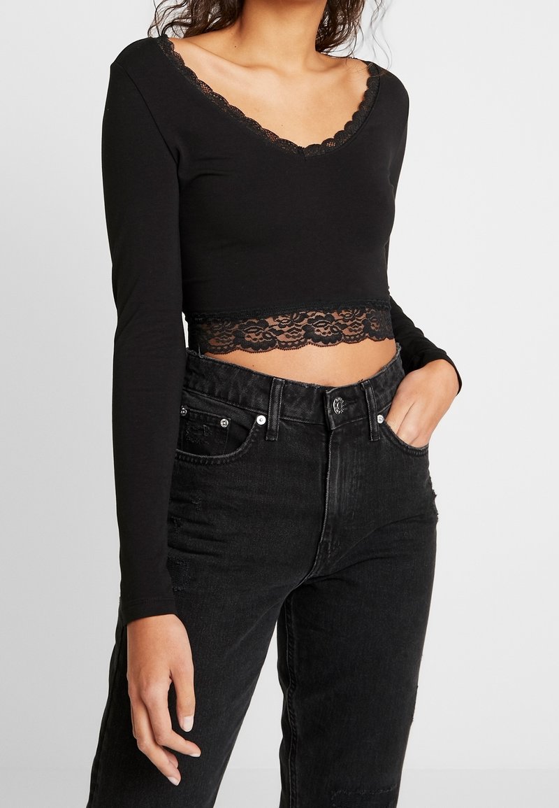 Frau in einem schwarzen langärmeligen Crop-Top mit Spitze und hochgeschnittenen schwarzen Jeans, Hand in der Tasche vor einem schlichten Hintergrund.