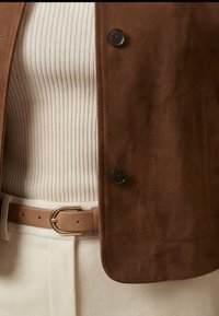 Veste en daim marron portée sur un haut blanc côtelé, pantalon beige, et ceinture en cuir camel avec boucle dorée.