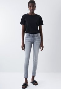 Graue enge Jeans mit hoher Taille und Fronttaschen, kombiniert mit einem schwarzen T-Shirt mit kurzen Ärmeln und schwarzen Rutschsandalen.