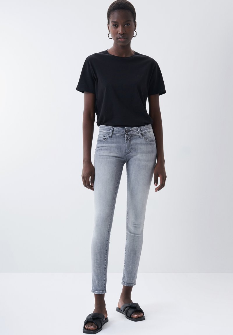 Graue enge Jeans mit hoher Taille und Fronttaschen, kombiniert mit einem schwarzen T-Shirt mit kurzen Ärmeln und schwarzen Rutschsandalen.