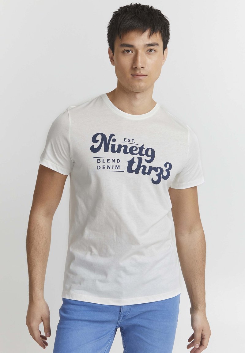 Bijela pamučna majica s okruglim izrezom i kratkim rukavima, s tamnoplavim natpisom "Ninetg thr33 Blend Denim" na prednjoj strani.