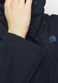 Main ajustant la capuche d'une veste The North Face bleu foncé avec logo visible et détails des boutons.