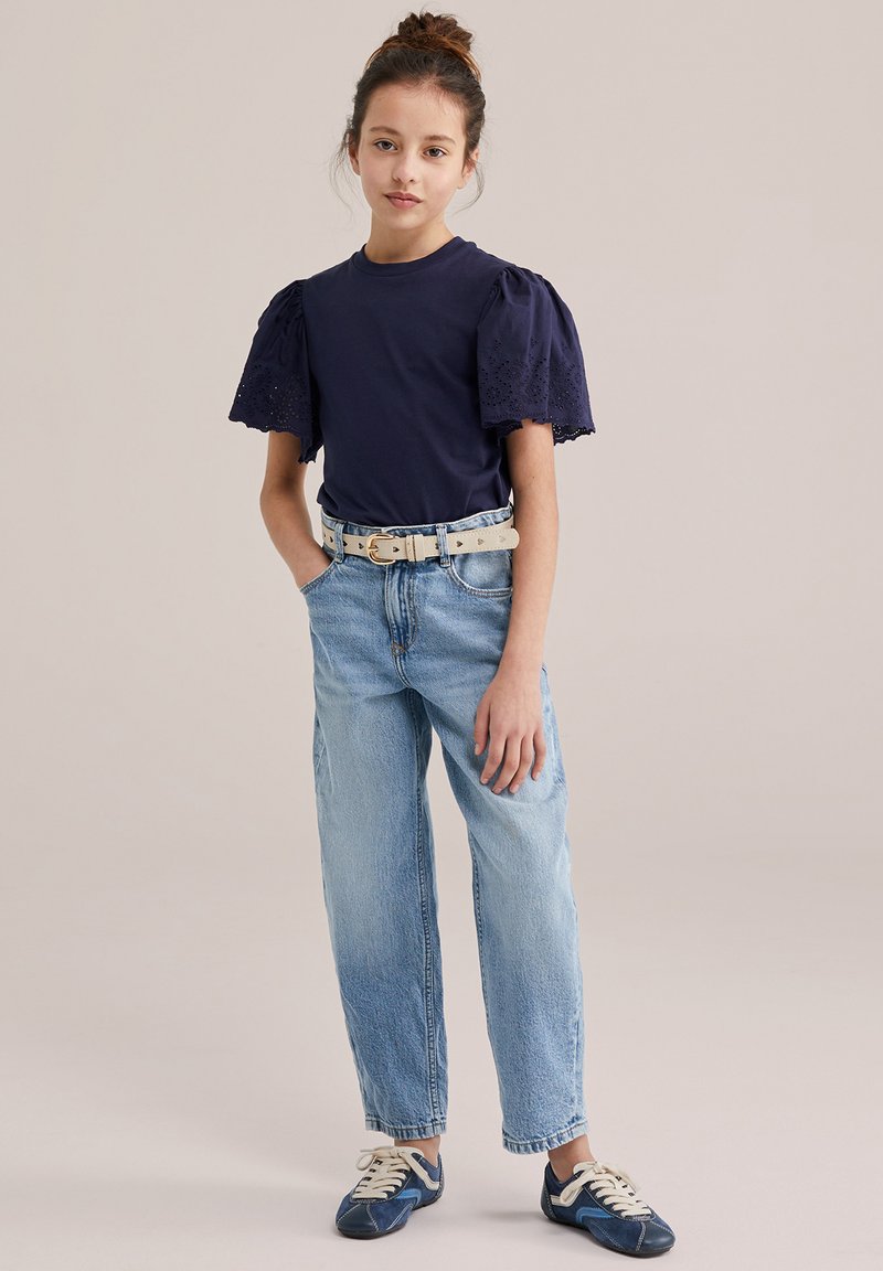 Jeune fille debout les mains dans les poches, portant un haut bleu marine à manches bouffantes, un jean bleu clair avec une ceinture beige, et des baskets bleues marines.