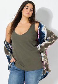 Frau mit langen dunklen Haaren, die ein olivgrünes Tanktop, blaue Jeans und ein teilweise abgenommenes Batikhemd trägt, steht vor einer weißen Wand.