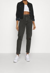 Person som bär en svart croppad denimjacka, en vit crop-top, högmidjade slitna svarta jeans och vita sneakers, stående mot en vit bakgrund.